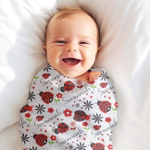 Pode incluir: Um cobertor de embrulho para beb&ecirc; branco com um padr&atilde;o repetido de joaninhas, flores e o nome "Savannah".