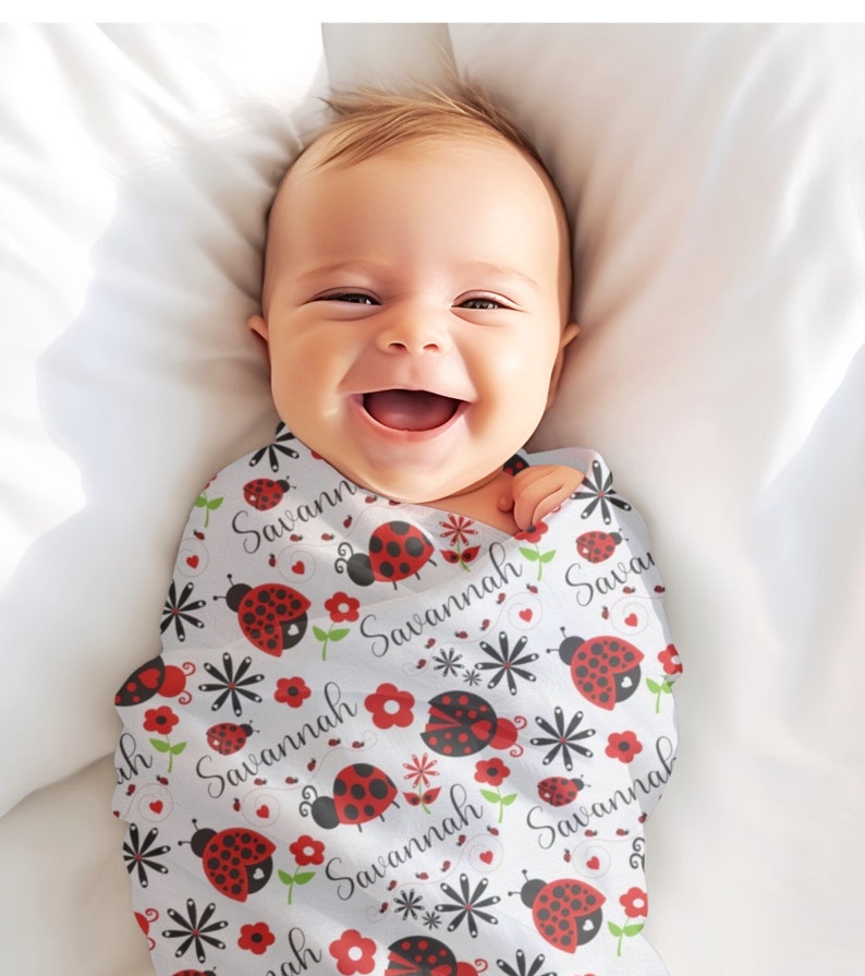 Pode incluir: Um cobertor de embrulho para beb&ecirc; branco com um padr&atilde;o repetido de joaninhas, flores e o nome "Savannah".