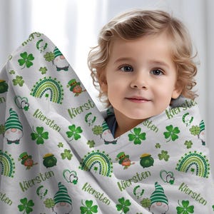 May include: A white and green personalized baby blanket with a shamrock, rainbow, and gnome pattern. The blanket has the name "Kiersten" repeated throughout the design.
