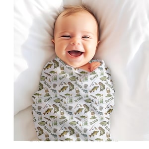 May include: A smiling baby wrapped in a white swaddle blanket. The blanket is patterned with illustrations of fish, fishing gear, and the name "OLIVER" in a repeating pattern. The baby is lying on a white surface.