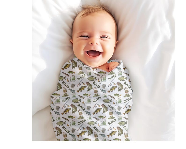 May include: A smiling baby wrapped in a white swaddle blanket. The blanket is patterned with illustrations of fish, fishing gear, and the name "OLIVER" in a repeating pattern. The baby is lying on a white surface.