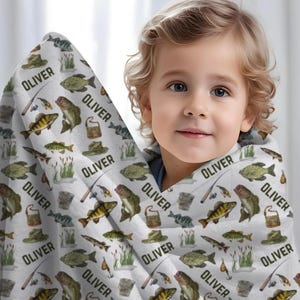 May include: A white blanket with a repeating pattern of fish, fishing hats, fishing poles, and the name "OLIVER" in black. The blanket is wrapped around a young child.