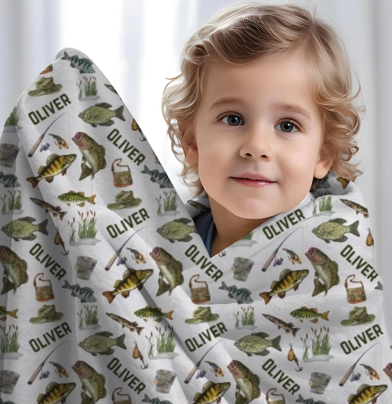 May include: A white blanket with a repeating pattern of fish, fishing hats, fishing poles, and the name "OLIVER" in black. The blanket is wrapped around a young child.