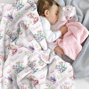 May include: A baby rests under a white blanket printed with pastel dragons, butterflies, and clouds. The name "PAULINE GRACE" is repeated across the blanket. A pink plush toy and a soft pink blanket are also visible.