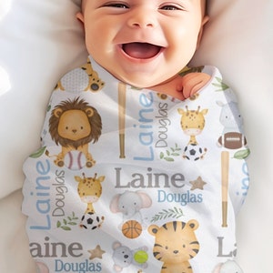 May include: A baby wrapped in a white swaddle blanket with a repeating pattern of cartoon animals, sports equipment, and the name "Laine Douglas" in blue and gray. The animals include a lion, giraffe, tiger, and elephant.