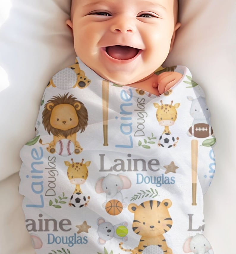 May include: A baby wrapped in a white swaddle blanket with a repeating pattern of cartoon animals, sports equipment, and the name "Laine Douglas" in blue and gray. The animals include a lion, giraffe, tiger, and elephant.