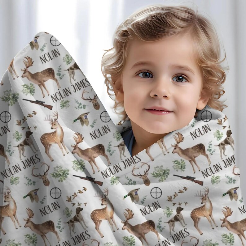 May include: A white blanket with a hunting theme, featuring deer, dogs, rifles, and the name "NOLAN JAMES" printed repeatedly. The blanket is wrapped around a child.
