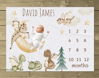 May include: A white baby blanket with a brown and yellow crescent moon and a brown dragon. There are four other dragons on the blanket, two green and two brown. The blanket also has a green pine tree and the text "DAVID JAMES" and "1 2 3 4 5 6 7 8 9 10 11 12 months".