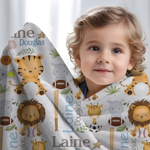 May include: A white baby blanket featuring cartoon animals like a tiger, lion, giraffe, and hippo, along with sports equipment. The blanket has the name "Laine Douglas" printed in blue and gray.