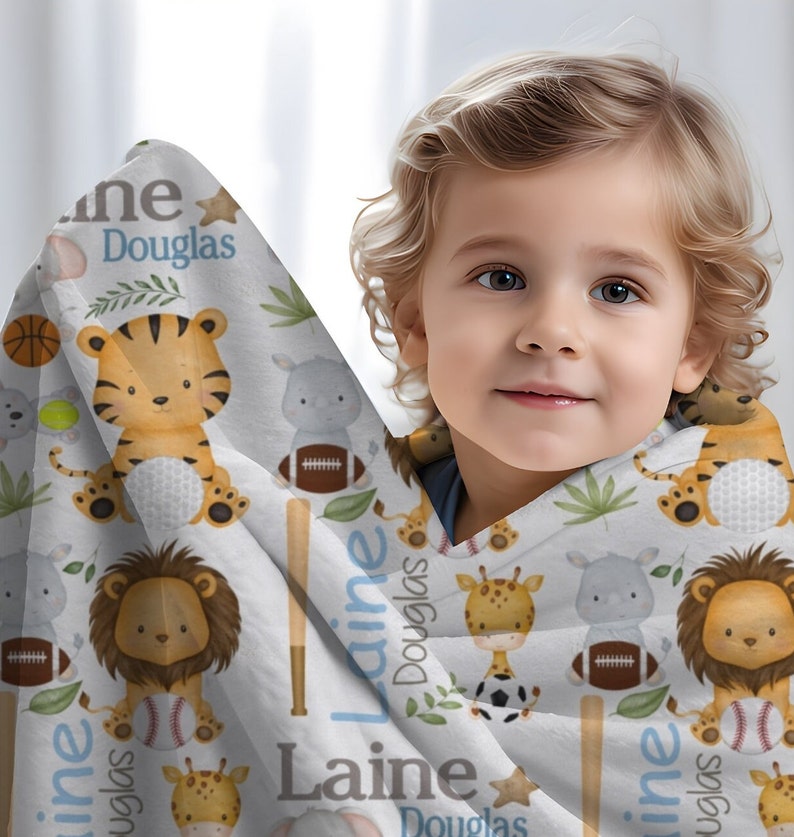 May include: A white baby blanket featuring cartoon animals like a tiger, lion, giraffe, and hippo, along with sports equipment. The blanket has the name "Laine Douglas" printed in blue and gray.