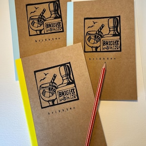 May include: Three notebooks with brown covers and light blue or yellow spines. Each notebook features a black and white illustration of a cocktail and the words "BRIGHT GIN" and "brighton". A red and white pencil rests on one of the notebooks.
