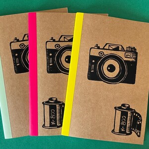 May include: Three rectangular notebooks with brown kraft paper covers, each featuring a black ink illustration of a camera and a film canister. The spines are accented with neon green, hot pink, and bright yellow tape. A black pencil lies nearby.