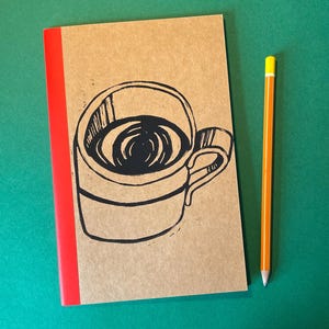 Op de afbeelding: Een beige notitieboek met een rode rug en een zwarte inktillustratie van een koffiemok op de voorkant, vergezeld van een geel en oranje potlood met een zilveren huls en een witte gum, verticaal naast het notitieboek geplaatst.