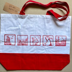 May include: A white cotton tote bag with red handles and a red bottom panel. The bag features five square red ink prints depicting a seaside theme, including a lighthouse, beach chairs, an ice cream cone, a seagull, and beach huts. A brown tag reads 100% cotton Hand wash only.