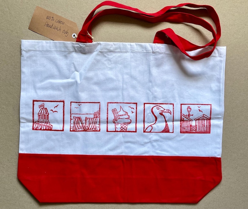 May include: A white cotton tote bag with red handles and a red bottom panel. The bag features five square red ink prints depicting a seaside theme, including a lighthouse, beach chairs, an ice cream cone, a seagull, and beach huts. A brown tag reads 100% cotton Hand wash only.