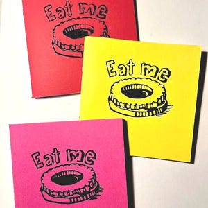 Peut inclure: Trois cartes colorées, rouge, jaune et rose, avec l'inscription "Eat me" sur une illustration en noir et blanc d'un biscuit. Les cartes sont disposées sur une surface blanche.