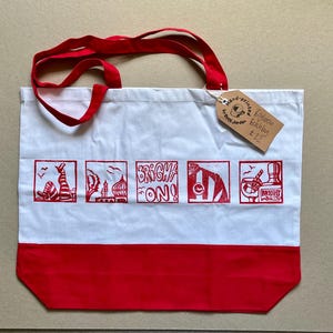 May include: A white tote bag with red handles and a red bottom panel. The bag features five red square illustrations depicting Brighton landmarks and the text "BRIGHT ON!". A brown cardboard tag is attached, reading "Hand-printed by Betti Jordan Brighton Beach bag &pound;22".