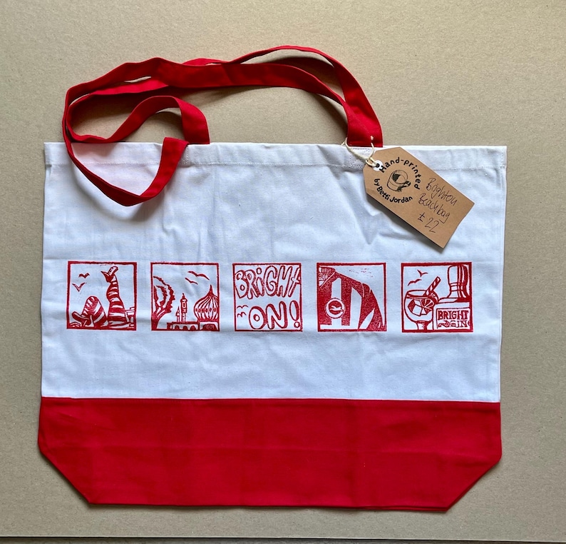 May include: A white tote bag with red handles and a red bottom panel. The bag features five red square illustrations depicting Brighton landmarks and the text "BRIGHT ON!". A brown cardboard tag is attached, reading "Hand-printed by Betti Jordan Brighton Beach bag &pound;22".