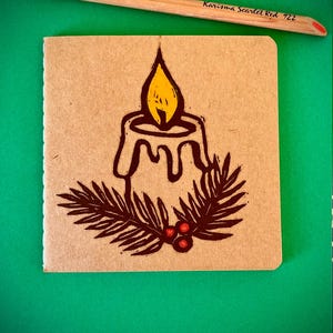 May include: A small, square notebook made of kraft paper, featuring a candle and evergreen branch design. A wooden pencil with "Karisma Scarlet Red 722" printed on it lies above the notebook on a green surface.