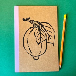 May include: A rectangular notebook with a tan cover featuring a black ink illustration of a lemon with leaves. The notebook has a light purple spine and is positioned next to a yellow wooden pencil with a silver ferrule and white eraser.