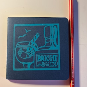 May include: A navy blue notebook with a teal illustration of a cocktail and the words "BRIGHT GIN." A red and white striped pencil is next to the notebook.