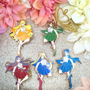 May include: Six enamel pins depicting anime characters in yellow, green, red, and blue. Each pin shows a character with long hair, a skirt, and a sword. The pins are set against a gold glitter background with floral accents.