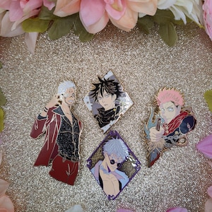 May include: Four enamel pins featuring anime characters. The pins depict characters in various poses and outfits, with vibrant colors and intricate details. The pins are set against a glittery gold background, surrounded by pink and white flowers.
