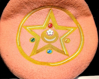 May include: A light peach-colored beret with a gold embroidered star design. The star is surrounded by a gold circle and adorned with small gemstones in red, green, and blue, and a white flower.