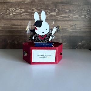 May include: A graduation-themed gift box featuring a white bunny wearing a graduation cap and gown. The box is red with a white label that reads "Happy Graduation! You did it!" The bunny holds a diploma and a banner that says "Way to go!"
