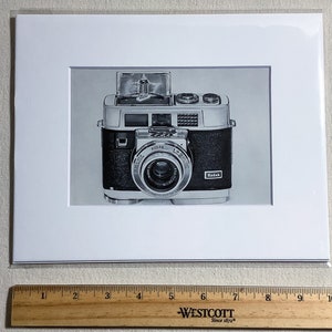 May include: Black and silver Kodak camera print, framed in white. The camera has a prominent lens and the Kodak logo is visible. The print is on a white mat, with a ruler at the bottom. The camera is a vintage model.