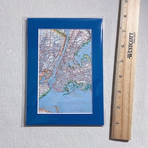May include: A framed map of New York City, featuring a blue frame. The map shows the city's layout with streets, landmarks, and water bodies. A wooden ruler is placed next to the frame, with measurements in inches.