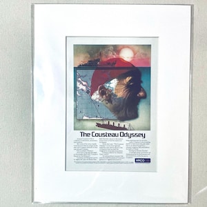 Op de afbeelding: Ingelijste print van de advertentie "The Cousteau Odyssey". Het kunstwerk toont een portret van Jacques Cousteau, een kaart, een schip en een rode zon. De tekst bevat de titel en details over het leven en de avonturen van Cousteau.