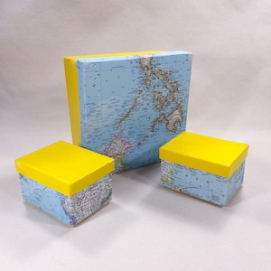 May include: Three decorative boxes with yellow lids and map-covered sides. The largest box is rectangular, and the other two are square. The boxes are arranged on a white surface.