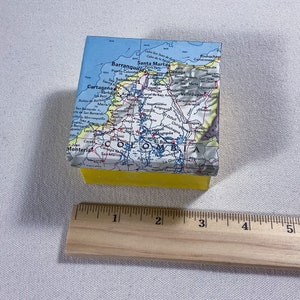 May include: A square soap bar with a map of Colombia on the top. The soap has a bright yellow base. A wooden ruler is placed next to the soap for scale. The soap measures approximately 5 cm.