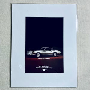 May include: A framed print featuring a 1977 Monte Carlo car advertisement. The car is white and silver, set against a dark background with the text "Like you, it's an original." and "Personal. Original. Affordable."
