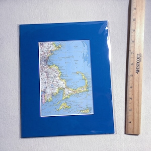 Op de afbeelding: Een ingelijste kaartafdruk van Cape Cod en Massachusetts, geplaatst tegen een levendige blauwe passe-partout. De kaart toont gedetailleerde wegennetwerken en geografische kenmerken. Een houten liniaal is rechts van de ingelijste afdruk geplaatst.