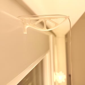 Könnte beinhalten: Ein klarer, geometrisch geformter Kunststoffclip zur Befestigung eines Drahtes, montiert auf einer weißen Oberfläche. Der Clip hat ein dreieckiges Design und ist an einer weißen Wand befestigt. Der Draht hängt herunter.