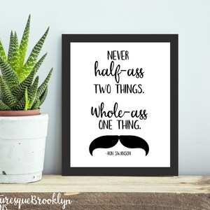 May include: A framed print with the quote "NEVER half-ass TWO THINGS. Whole-ass ONE THING." and a mustache graphic. A potted succulent plant sits to the left. The print is on a wooden shelf.