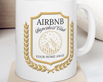 May include: White ceramic mug with the text "AIRBNB Superhost Club" in black and gold. The mug features a shield design with a mountain and tree graphic. The mug is on a beige coaster.
