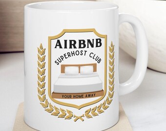 May include: White ceramic mug with a gold crest featuring the text "Airbnb Superhost Club" and a bed with two pillows. The crest is surrounded by a laurel wreath and the text "Your Home Away" is below the bed.