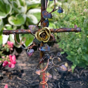 May include: A metal garden rain gauge with a copper finish and a gold rose design. The gauge is shaped like a cross with a vine design and is surrounded by green foliage.