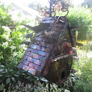 Puede incluir: Una pajarera decorativa hecha a mano con un techo de metal iridiscente multicolor. La pajarera tiene un agujero de entrada redondo, detalles decorativos de metal y una flor roja. Est&aacute; montada sobre un soporte de metal y rodeada de vegetaci&oacute;n. Mide unos 30 cm de alto.