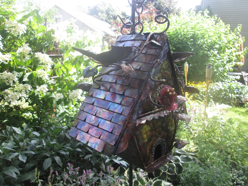 Puede incluir: Una pajarera decorativa hecha a mano con un techo de metal iridiscente multicolor. La pajarera tiene un agujero de entrada redondo, detalles decorativos de metal y una flor roja. Est&aacute; montada sobre un soporte de metal y rodeada de vegetaci&oacute;n. Mide unos 30 cm de alto.