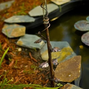 May include: A metal rain gauge with a decorative vine design and leaves. The gauge is placed in a garden setting with rocks and mulch.