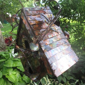 Puede incluir: Una pajarera de metal hecha a mano con un tejado color cobre y detalles intrincados de flores y ramas. La pajarera tiene dos agujeros de entrada circulares y est&aacute; suspendida de una cadena de metal. El dise&ntilde;o general es r&uacute;stico y decorativo.