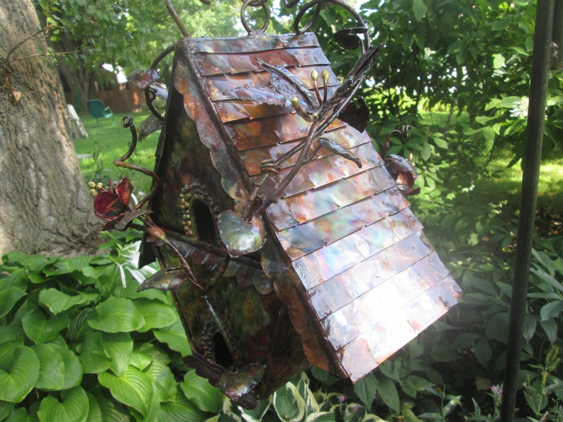Puede incluir: Una pajarera de metal hecha a mano con un tejado color cobre y detalles intrincados de flores y ramas. La pajarera tiene dos agujeros de entrada circulares y est&aacute; suspendida de una cadena de metal. El dise&ntilde;o general es r&uacute;stico y decorativo.