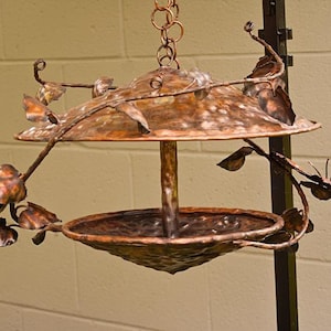 May include: A handcrafted copper bird feeder with a decorative leaf and branch design. The feeder has a circular top and a bowl-shaped base, suspended by a chain. The copper has a natural, aged patina, giving it a rustic appearance.