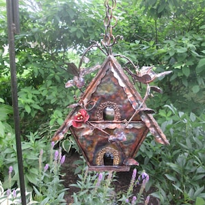 Puede incluir: Una pajarera decorativa de metal con un acabado r&uacute;stico multicolor. La pajarera presenta dos aberturas, p&aacute;jaros decorativos y detalles florales. Est&aacute; suspendida por una cadena y se encuentra sobre un fondo de exuberante follaje verde.