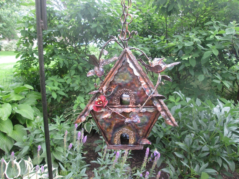 Puede incluir: Una pajarera decorativa de metal con un acabado r&uacute;stico multicolor. La pajarera presenta dos aberturas, p&aacute;jaros decorativos y detalles florales. Est&aacute; suspendida por una cadena y se encuentra sobre un fondo de exuberante follaje verde.
