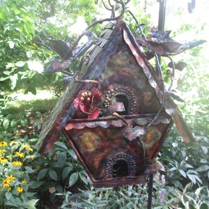Puede incluir: Una pajarera de metal hecha a mano con un aspecto r&uacute;stico y envejecido. La pajarera presenta un tejado a dos aguas, dos orificios de entrada circulares y detalles decorativos de flores y hojas. La paleta de colores incluye tonos de marr&oacute;n, cobre y rojo, con un toque de verde.